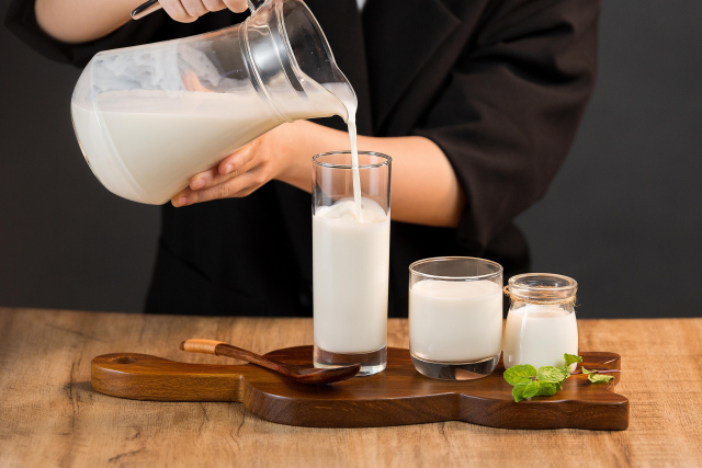 ピッチャーからグラスに牛乳を注いでいる様子。木製のトレイの上には、他にも牛乳が入ったグラスが２つ置かれている