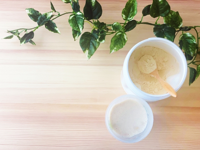 木のテーブルの上に置かれた、粉末プロテインの容器と、それを溶かして作ったプロテインドリンク、そして観葉植物