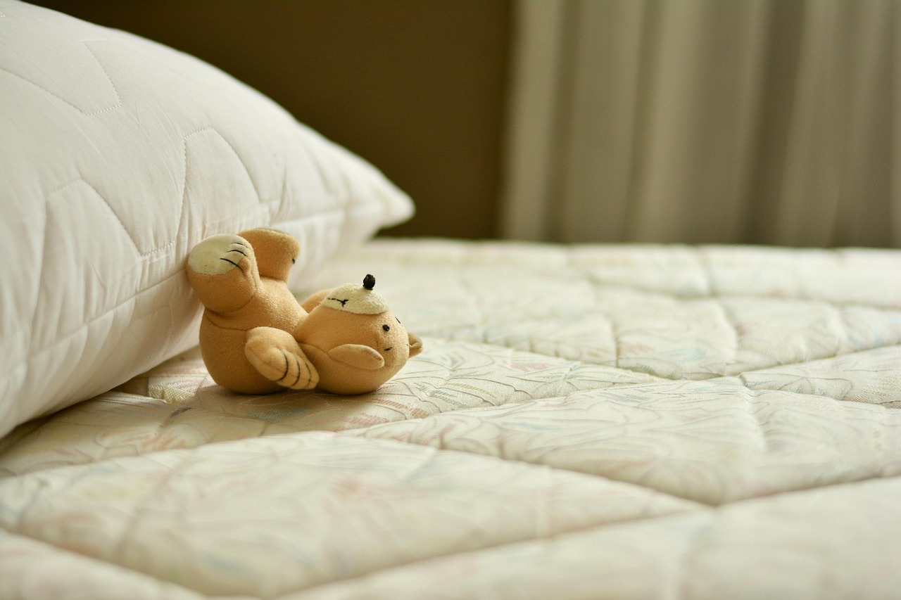 ベッドの上に置かれた白い枕と、その横で仰向けになって寝ているような、小さな茶色のテディベアのぬいぐるみ。