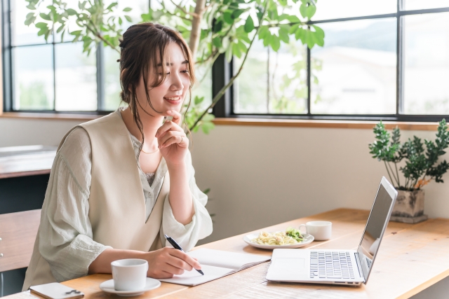 明るいカフェでノートパソコンを使い、ペンを持って考え事をする笑顔の女性