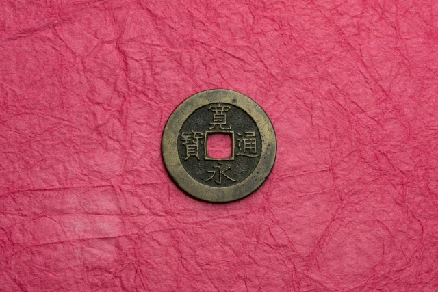 赤色の和紙のような背景の上に置かれた、江戸時代の古銭「寛永通宝（かんえいつうほう）」。中央に正方形の穴があり、上下左右に「寛永通宝」の文字が刻まれている。