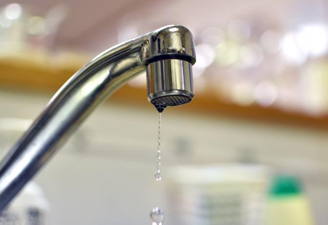 ステンレス製の蛇口の先端から、一滴の水が滴り落ちている様子。背景はぼかされた室内のデスクや物が見える。